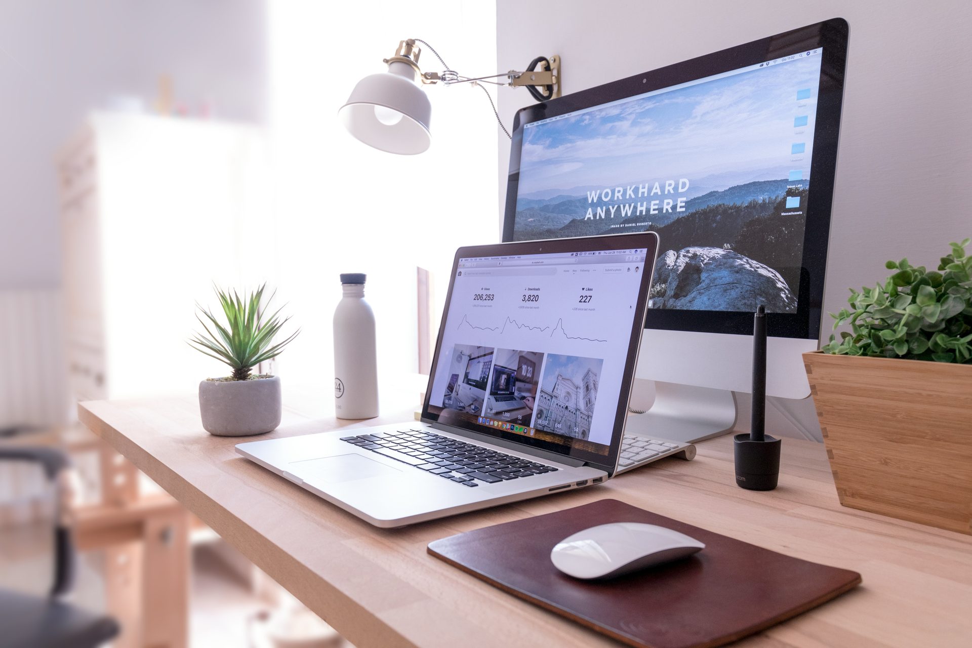 A laptop and desktop on a bright, plant-filled desk. The kind of workspace a remote-first role makes possible.
